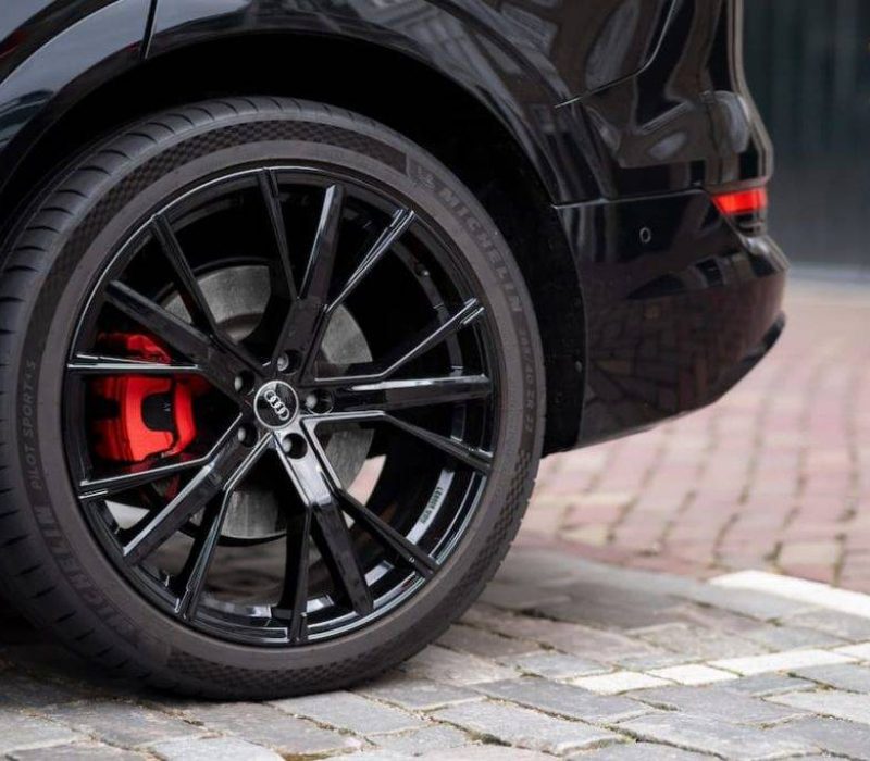 Achterwiel van een zwarte Audi met glanzend zwarte velg en donkerblauwe remklauw, geparkeerd op een geplaveide straat.